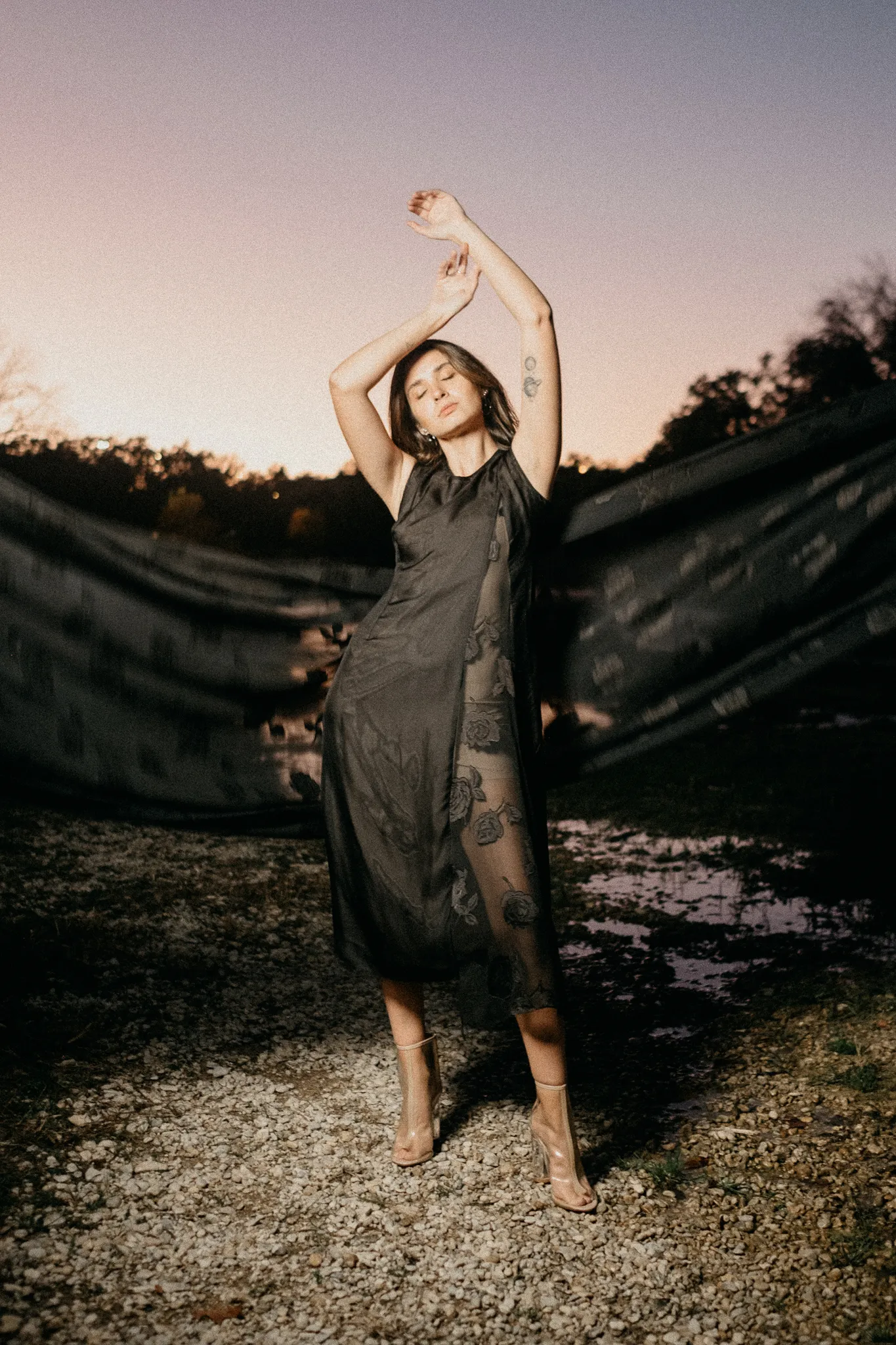 Model posing outdoors at sunset wearing a Looking Friday black sheer floral silk sleeveless midi dress