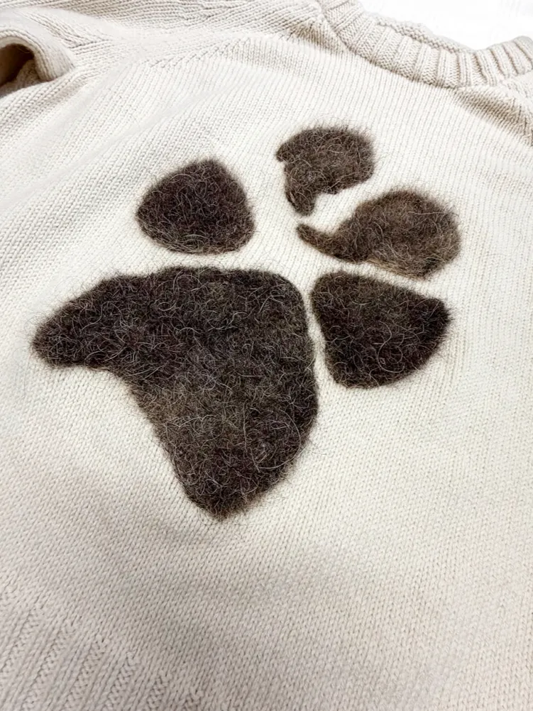 A close-up view of a cream-colored, textured knitted sweater featuring a large, dark brown, fuzzy felted paw print appliqué centered on the chest.