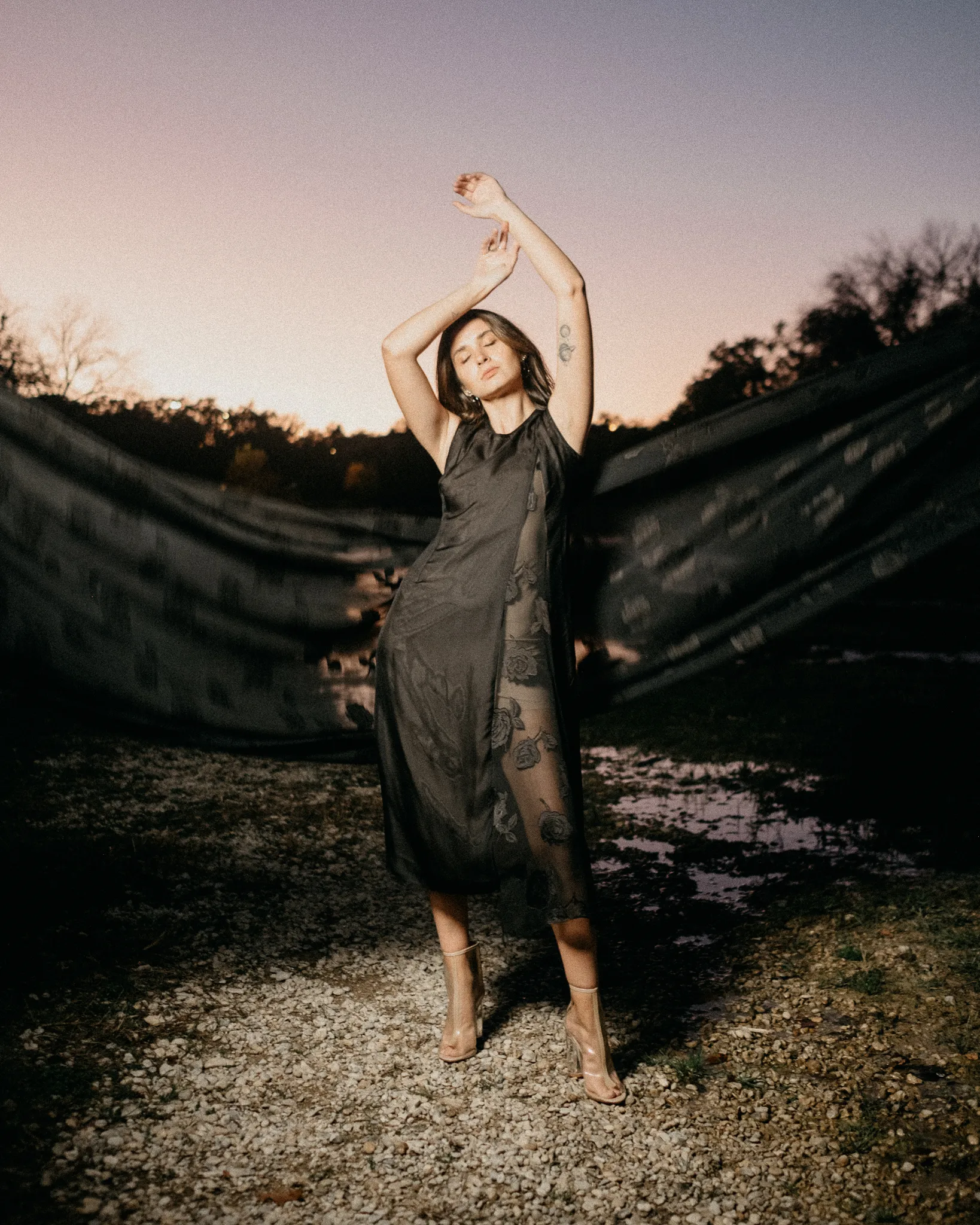 Model posing at twilight in Looking Friday black sleeveless dress with sheer floral rose jacquard silk panels.