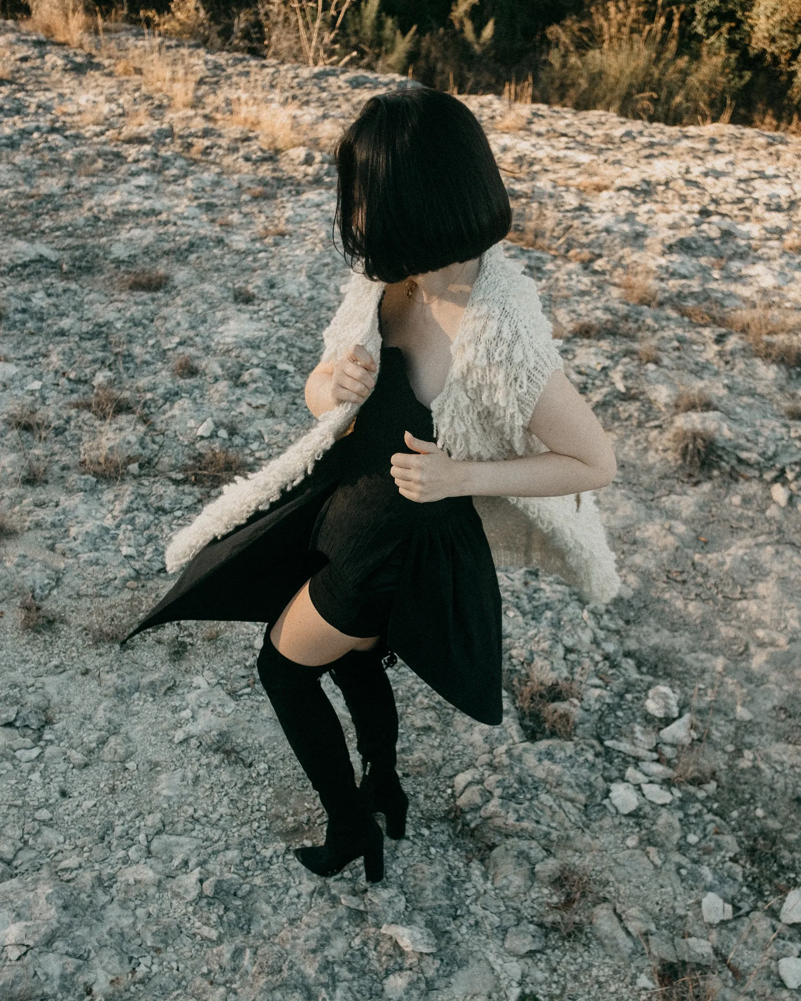 Model on rocky terrain in Looking Friday cream loopy stitch cardigan vest over black heavy-weight cotton pleated corset top.