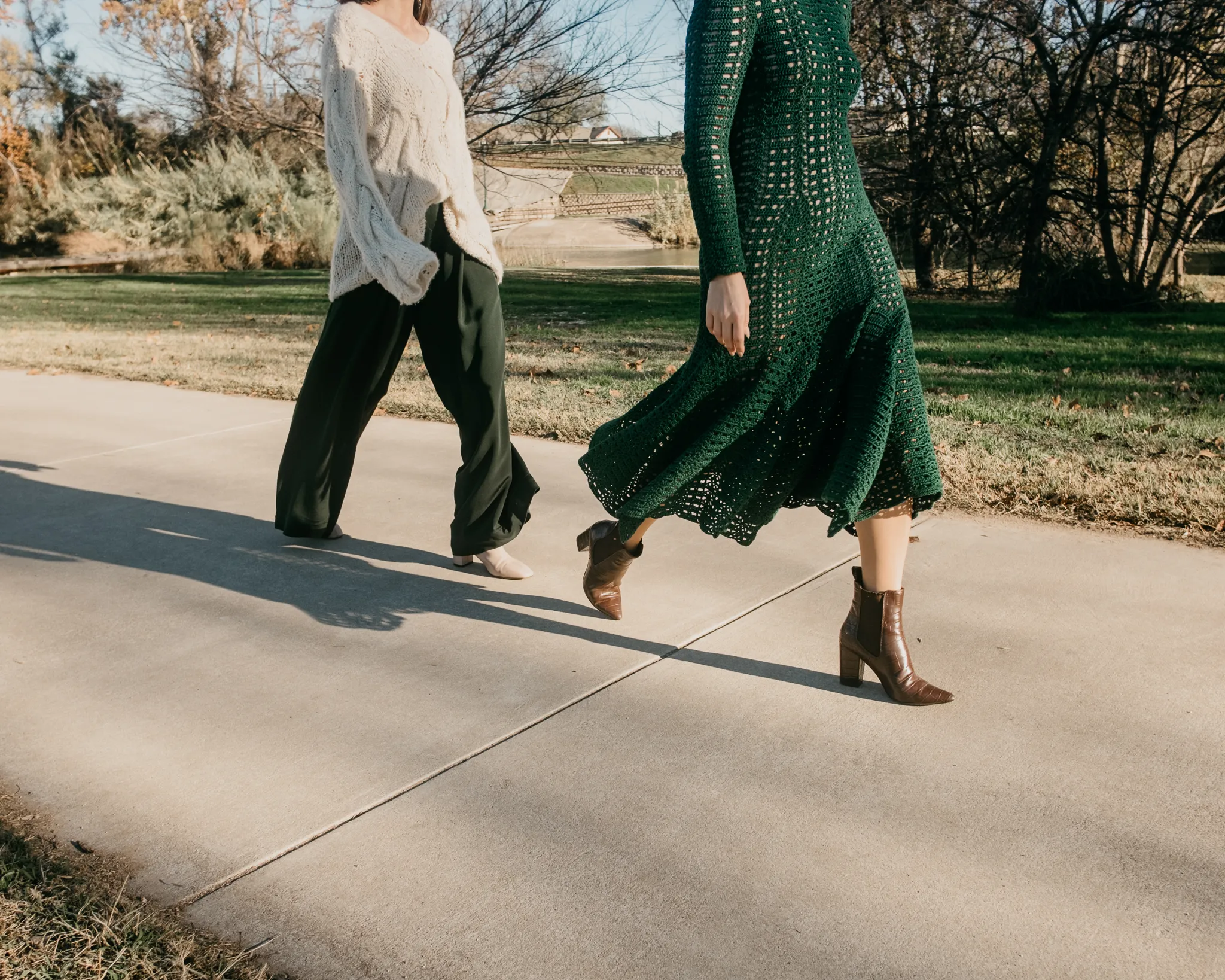 Two models walking outdoors in Looking Friday green hand-crocheted lattice dress, cream oversized cable cashmere sweater, and dark green raw silk trousers.