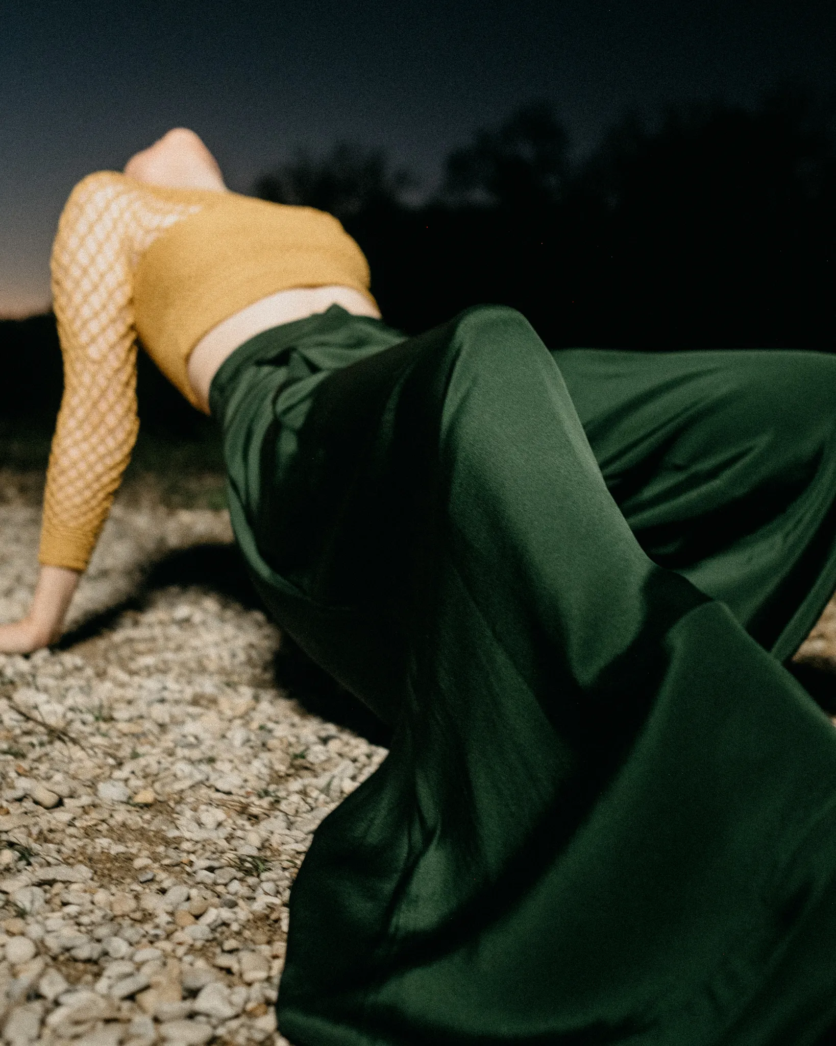 Model leaning back on gravel wearing Looking Friday forest green 100% raw silk wide-leg Executive Pants and mustard yellow crochet mesh cropped top.