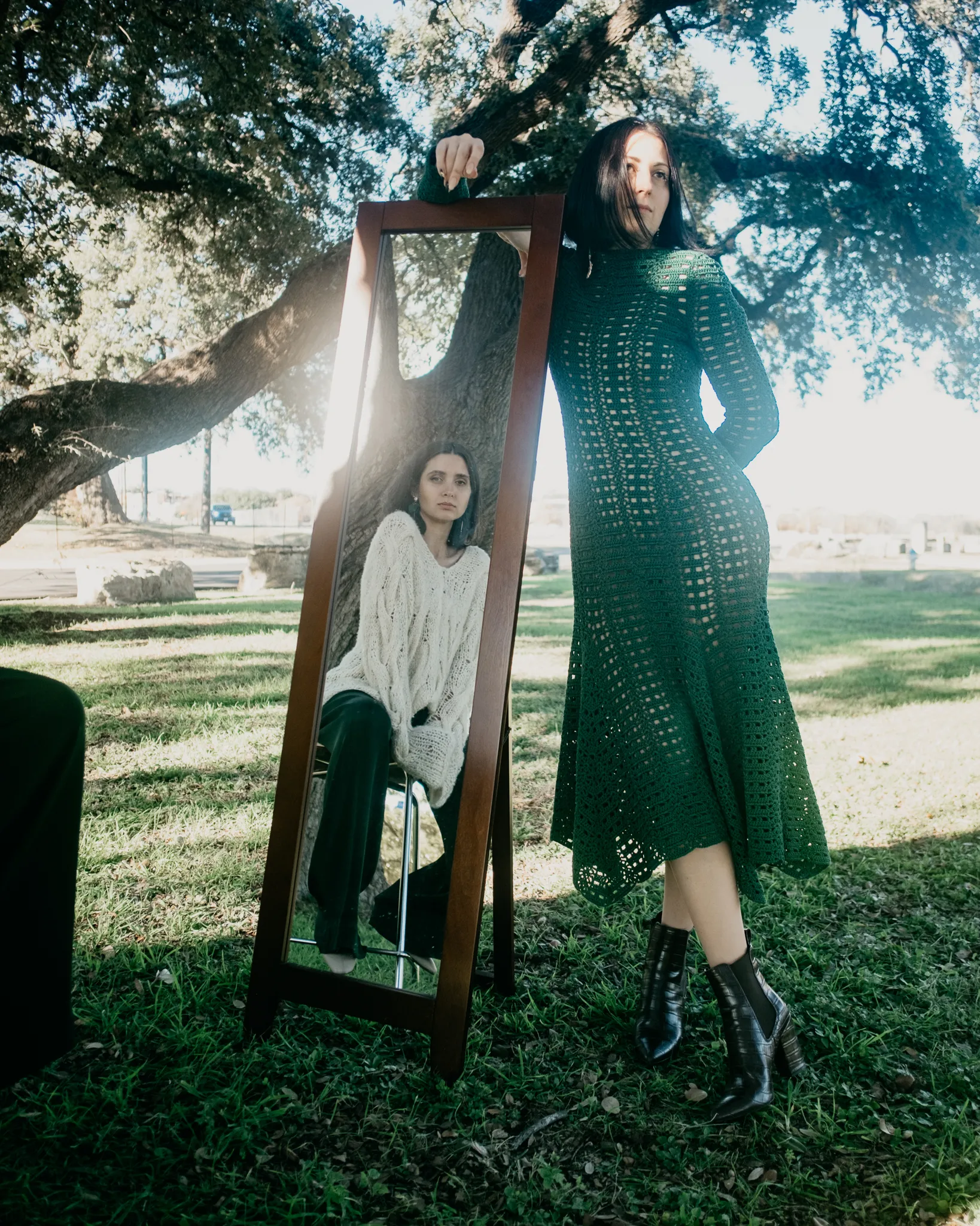 Model in Looking Friday forest green Italian cotton crochet lace dress posing outdoors beside a mirror reflecting a cream oversized cable knit sweater.