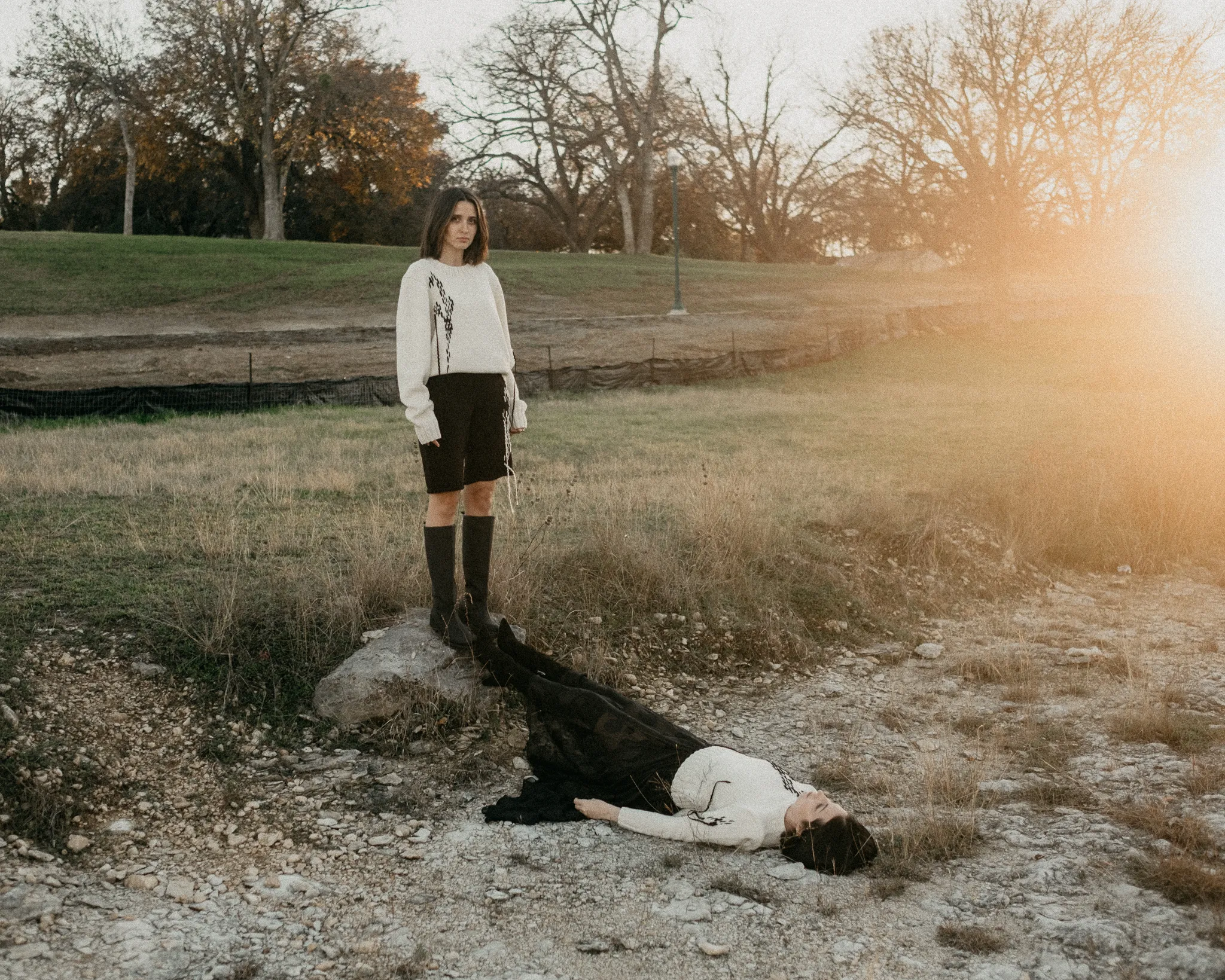 Two models in a field at sunset; one standing in a white merino wool sweater and knit shorts, the other lying down in a black silk rose jacquard skirt.