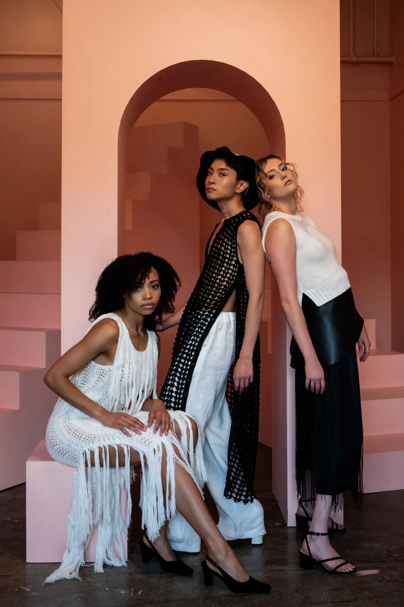 Group of models in Looking Friday designs: white Hand-Knitted Fringe Dress, black Crochet Fan Tunic with white trousers, and Distressed Knit Top with black culottes.