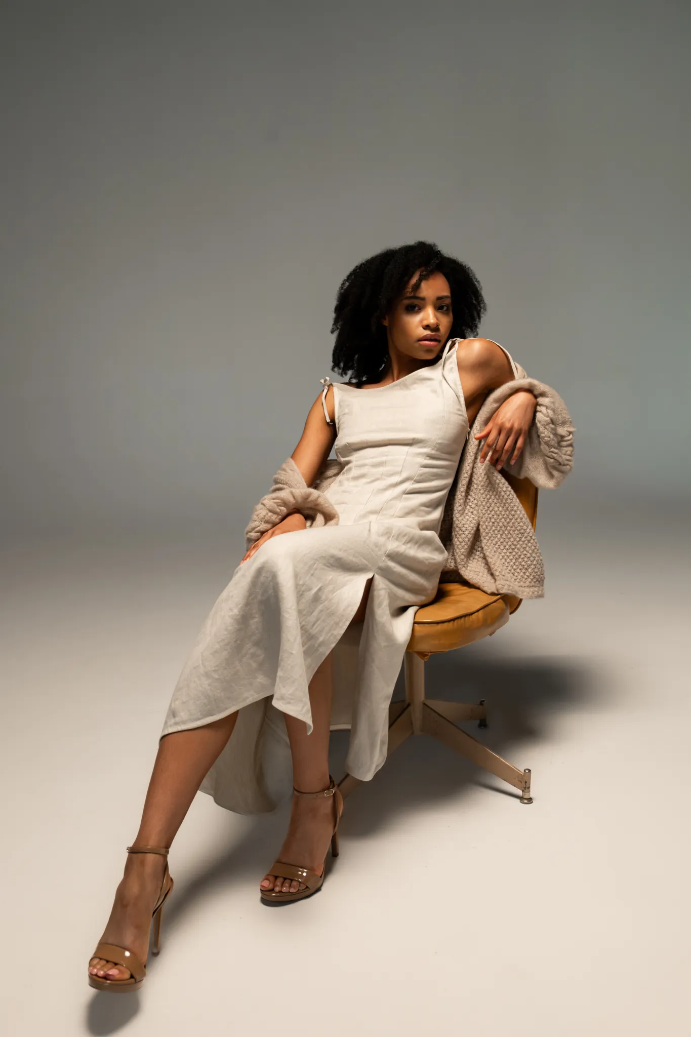 Model seated in Looking Friday natural beige Linen Summer Dress with shoulder ties paired with an oversized Hand-Knitted Cashmere Cardigan.