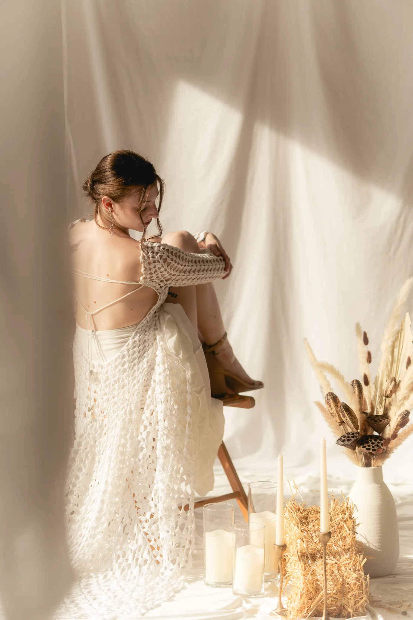 Seated bride wearing the Naiad Gown by Looking Friday, featuring a low back, hand-crocheted cotton texture, and soft silk lining drape in a warm studio setting.