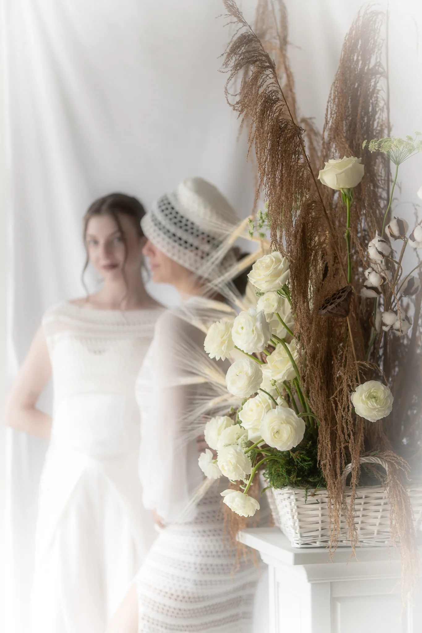 Soft-focus editorial portrait showing a bride wearing Looking Friday's Sophie Midi Dress in hand-crocheted organic cotton with sheer silk sleeves and a pillbox hat, framed by white floral arrangements.